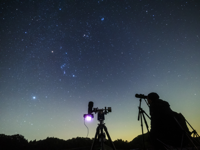 今回は視界の広いR1駐車場に赤道儀をセットして星雲や星団の撮影をした。画面中央の機材が望遠レンズでプレアデス星団を追尾撮影している赤道儀だ。右はイスに腰掛けてカメラ三脚に固定した口径70mm14倍の大型双眼鏡でオリオン大星雲を観望中の取材スタッフ。センターポールの傾けられる三脚だと双眼鏡が覗きやすくて楽チン。<br>OLYMPUS OM-D E-M5 Mark II/M.ZUIKO DIGITAL ED 7-14mm F2.8 PRO/ISO3200/7mm/F2.8開放/30秒/LEEソフトフィルターNo.3使用