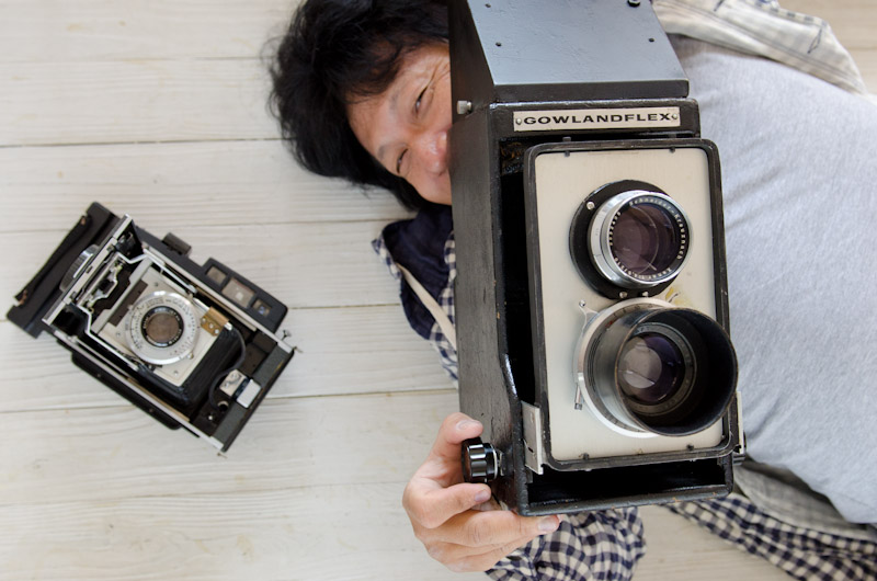 <b>70~80年代に流行った懐かしいジュラルミンケースにギッシリ詰まっているのはこれまた懐かしい往年のニコン、オリンパス、ペンタックス等の銀塩35㎜一眼レフカメラたち(左)。フジPETなどもあり、これまた懐かしい。右写真、幹幸くんが抱きかかえているのはぬいぐるみではなく、まぎれもなくホンモノのカメラである。GOWLANDFLEXというアメリカ製の4×5の手持ち撮影用巨大カメラ。これらの他にもクラシックカメラがもっとあるらしく見せたがっていたが、誌面(サイト?)の都合上これ以上は丁寧に遠慮させていただきました(笑)。</b>