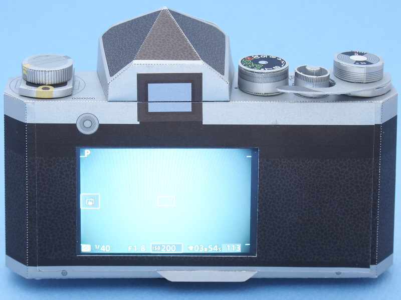 一見すると、フィルム一眼レフの名機「ニコンF」なのだが、実は中身は最新のデジカメで、もちろん撮影可能である。外装はホンモノのニコンFではなく、ニコンダイレクト限定グッズの「ニコンFペーパークラフト」で、つまり素材は紙である。その紙のニコンFに、ホンモノのデジカメ「Nikon1 V3」を内蔵し、さらに電源スイッチやシャッターボタンを操作する機構も組み込んだ。これらのパーツ類も、今回は全て紙で製作した。レンズは「1 NIKKOR 18.5mm f/1.8」を装着し、ニコンFペーパークラフトのレンズNikkor 50mm F1.4と同様の画角で撮影できる。